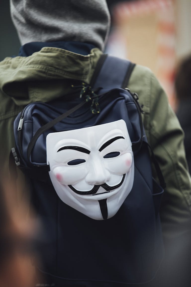 A person wearing a hoodie with a Guy Fawkes mask attached to a backpack, depicting anonymity.