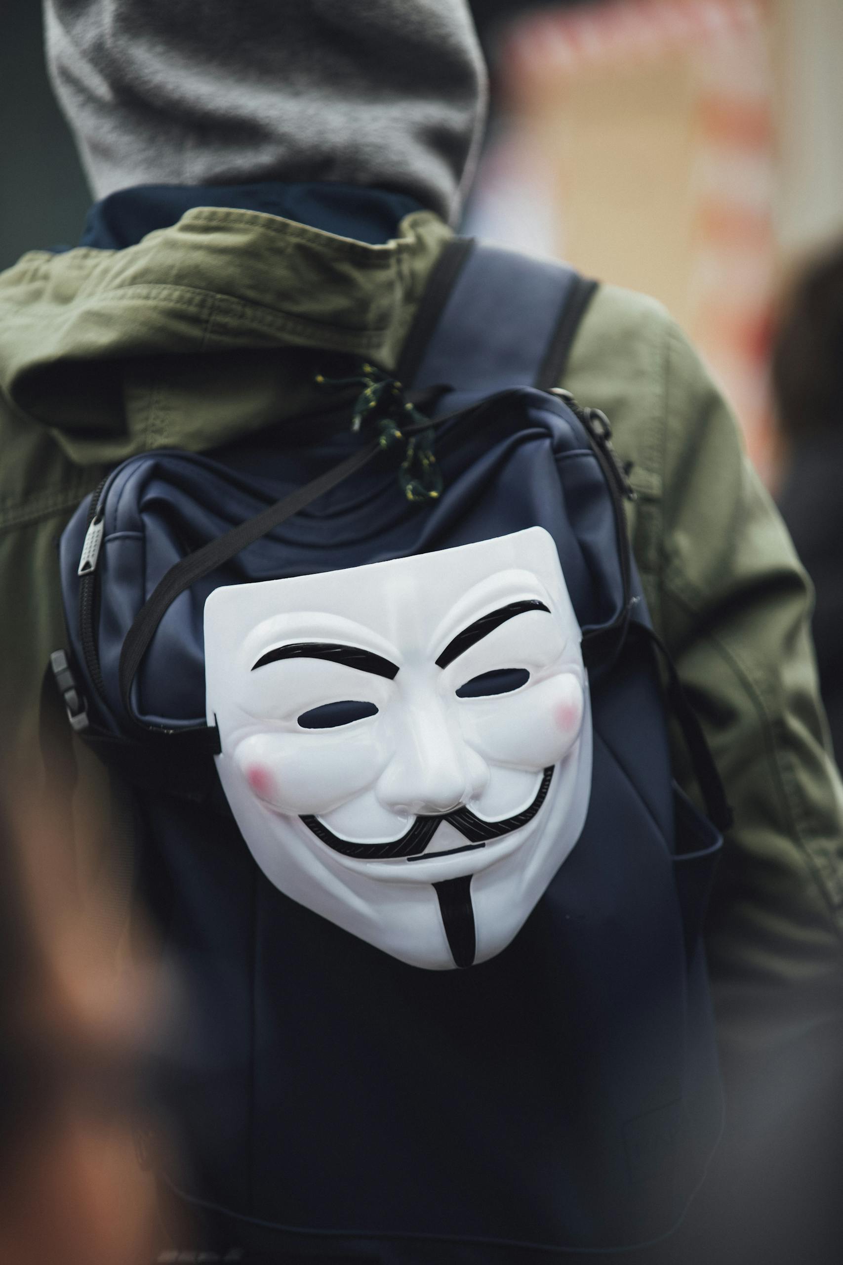 A person wearing a hoodie with a Guy Fawkes mask attached to a backpack, depicting anonymity.
